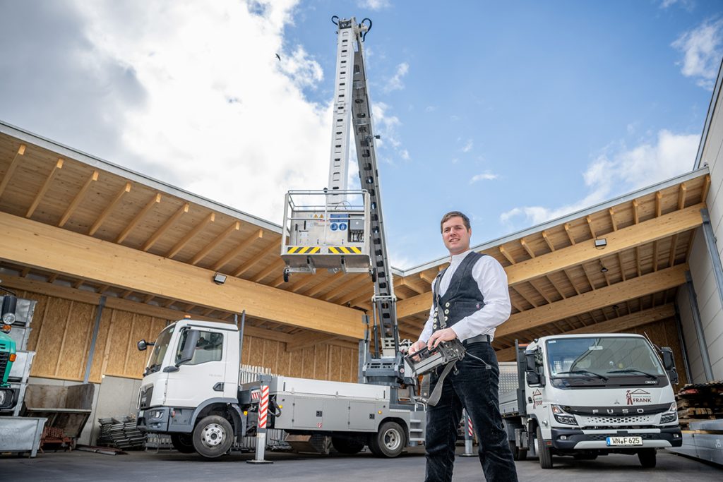 Geschäftsführer Martin Frank steht im Betriebshof der Firma, hinter ihm der Autokran der Firma mit ausgefahrener Hebebühne und ein weiteres Firmenfahrzeug.