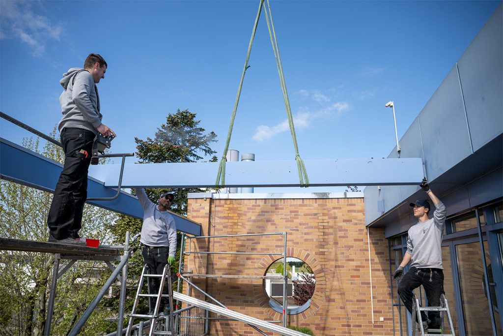 Geschäftsführer Martin Frank und zwei Mitarbeiter platzieren auf einer Dachterrasse einen großen Holzbalken, der an einem Kran hängt.