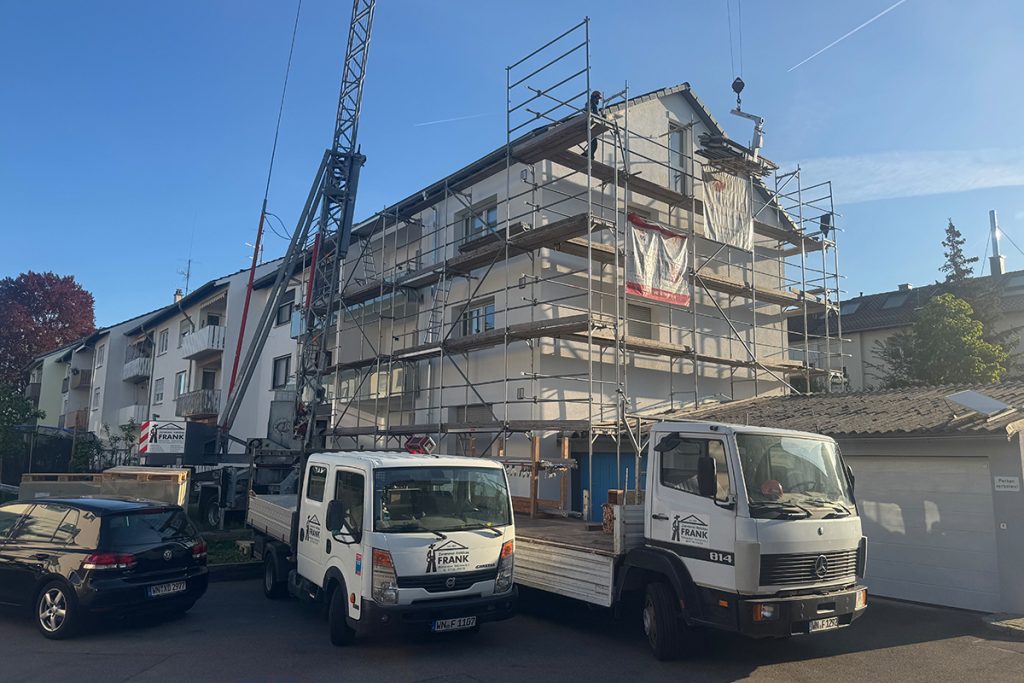 Ein Mehrfamilienhaus mit Baugerüst, davor stehen zwei Firmenfahrzeuge von Frank Zimmerei und Holzbau.